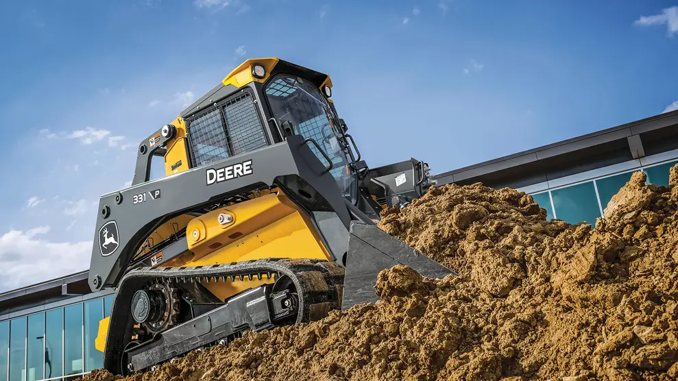 Yellow John Deere 331 P compact track loader on a dirt pile in front of a commercial building